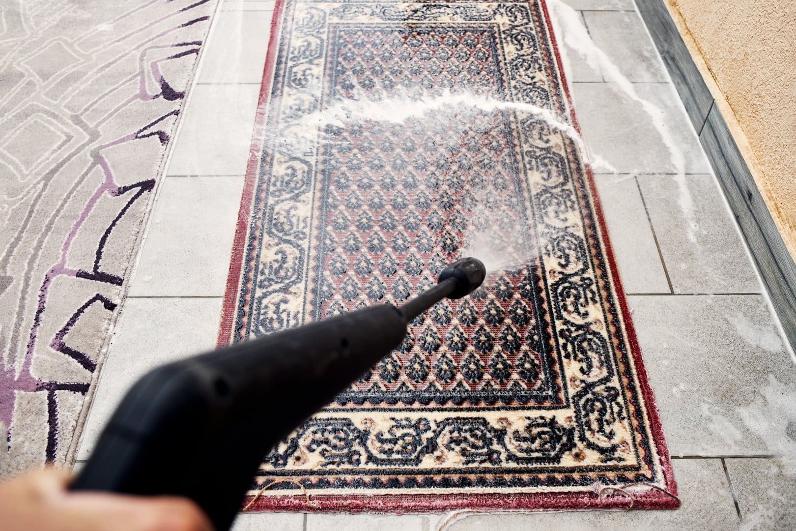 A dynamic top-down shot showing a traditional patterned rug being thoroughly cleaned on a tiled patio with a high-pressure washer.