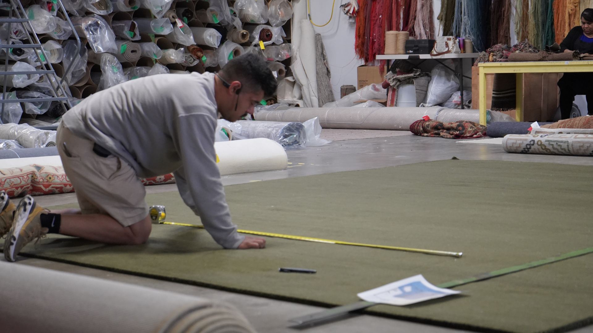 Rug specialist measuring a rug with a tape measure in a workshop lined with rolled Oriental rugs