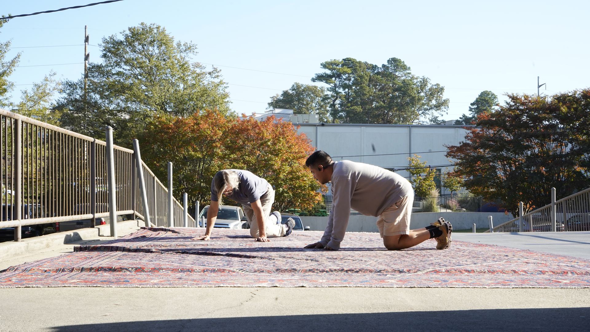 Two S&S Rug Cleaners technicians kneeling on a red hand-knotted Oriental rug outdoors to inspect its pile, wear, and condition